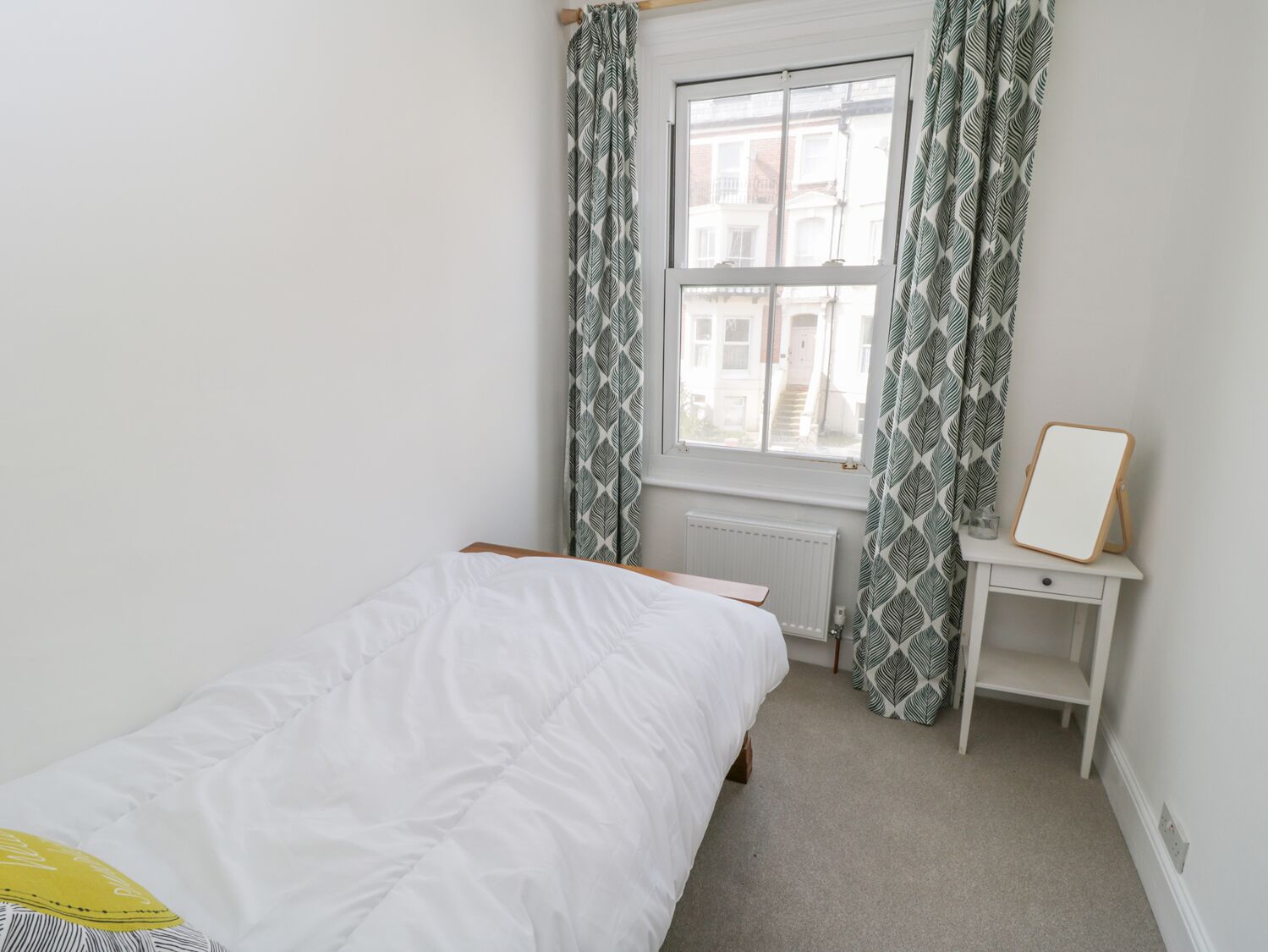 Une chambre avec un lit simple, une fenêtre avec des rideaux à motifs, une table de chevet avec un miroir et un radiateur.