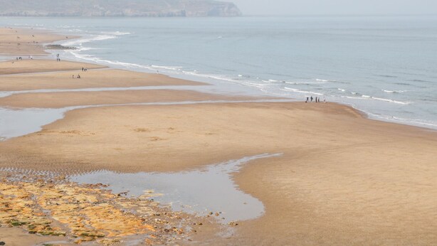 A wide sandy beach with gentle waves, a rocky shoreline, and distant cliffs. - West Cliff House (Whitby)