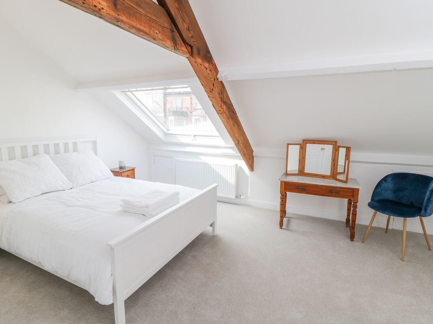 Ein Schlafzimmer mit einem weißen Bett, einem Holztisch, einem blauen Stuhl und einem Dachfenster.