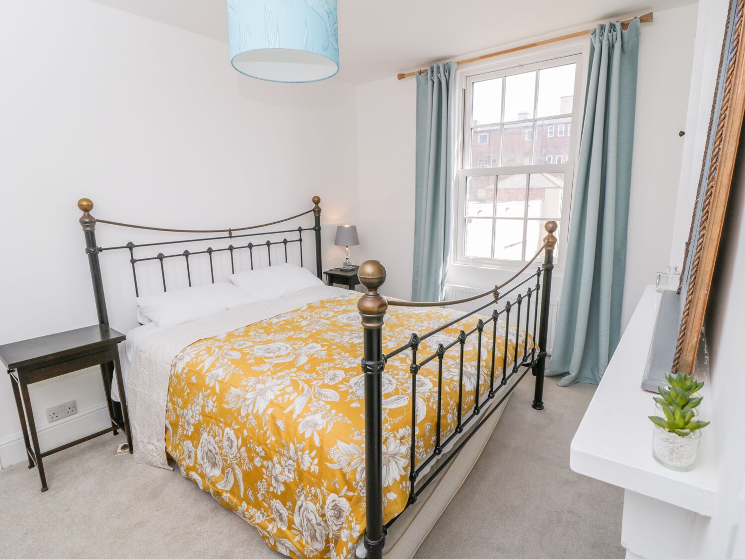 Une chambre à coucher avec un lit, une table de chevet, une lampe, une fenêtre avec des rideaux et une petite plante posée sur une étagère blanche.