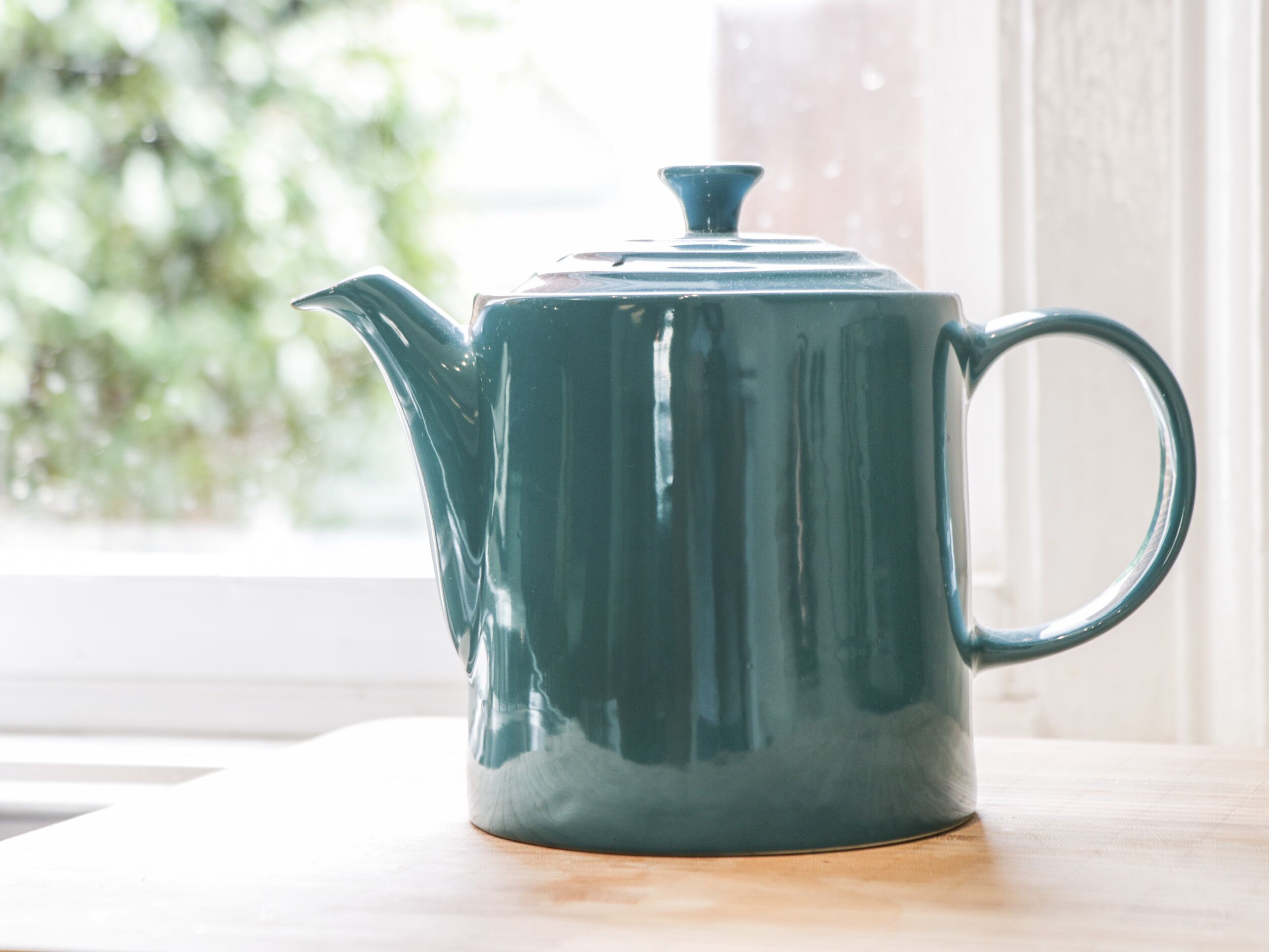 A teal ceramic teapot on a wooden surface near a window with a view of greenery.