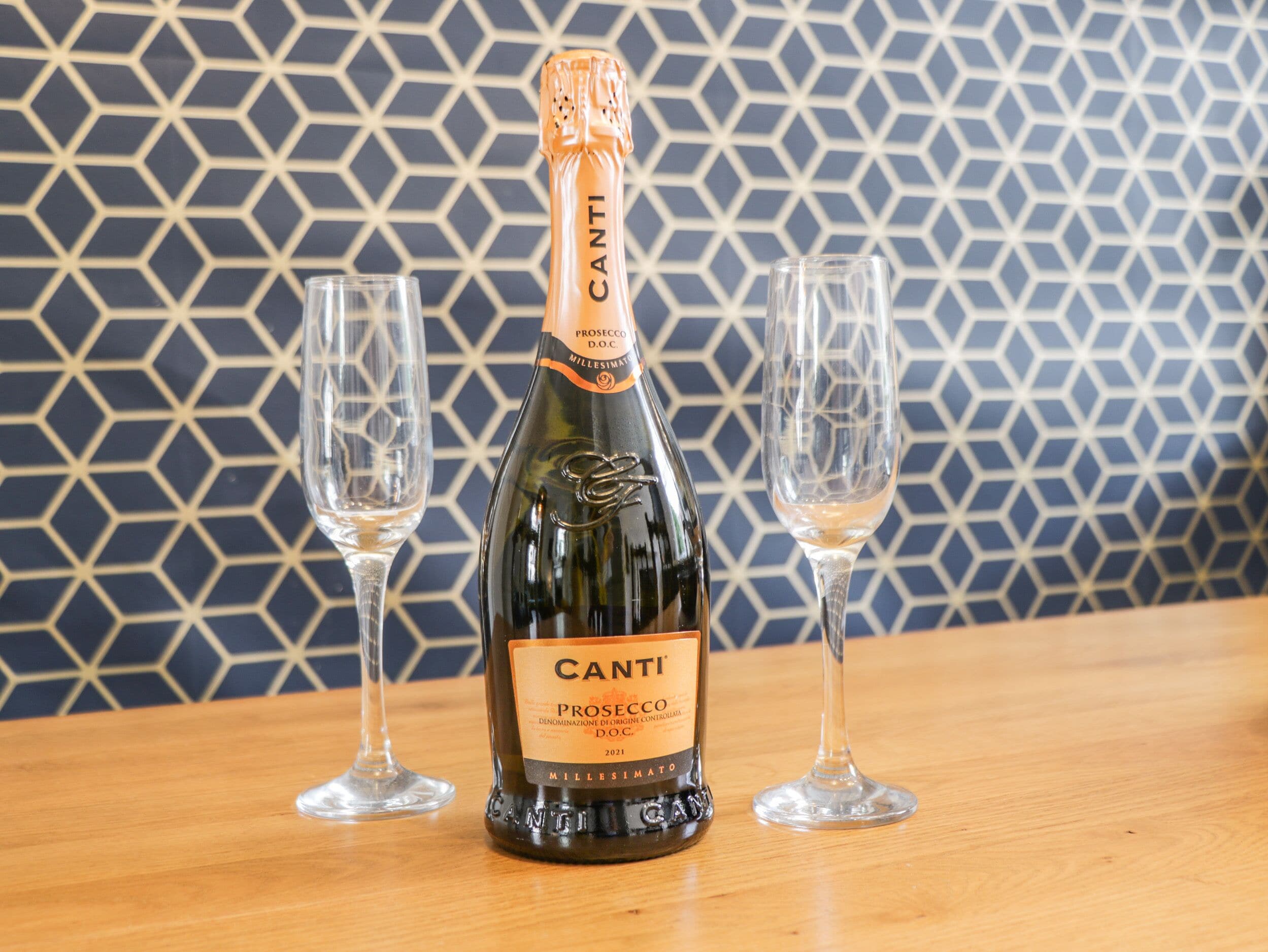 A bottle of Canti Prosecco D.O.C. 2021 on a wooden table with two empty wine glasses.