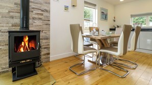 Un salon moderne doté d’un poêle à bois, un coin repas avec une table en bois et des chaises en métal, et une cuisine avec une fenêtre.