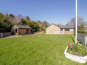 A stone building with a sloped roof, a small wooden structure with a glass enclosure, and a well-maintained lawn with a flower bed. - Cornflower Cottage (St. Columb Major)