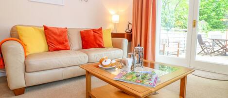 A living room with a beige sofa, orange cushions, a wooden coffee table with a magazine, a coffee pot, and a window with curtains.