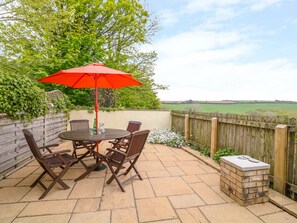 A patio with a round table and four chairs, a red umbrella, and a view of a green field. - Cornflower Cottage (St. Columb Major)