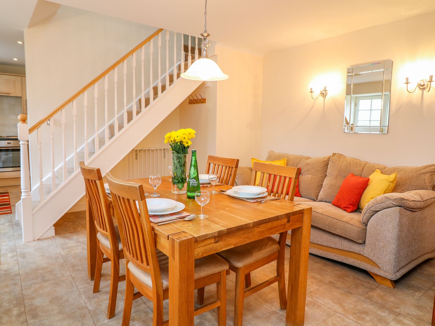 A dining area with a wooden table, chairs, and a vase of yellow flowers. A staircase is visible in the background.
