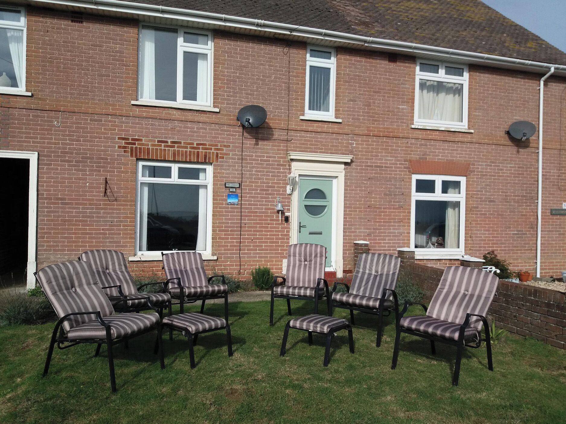 A brick house with a front door, a satellite dish, and a lawn with six striped chairs.