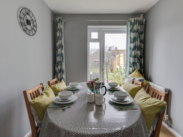 A dining area with a round table set for four, surrounded by wooden chairs with cushions. A clock is mounted on the wall above the table.