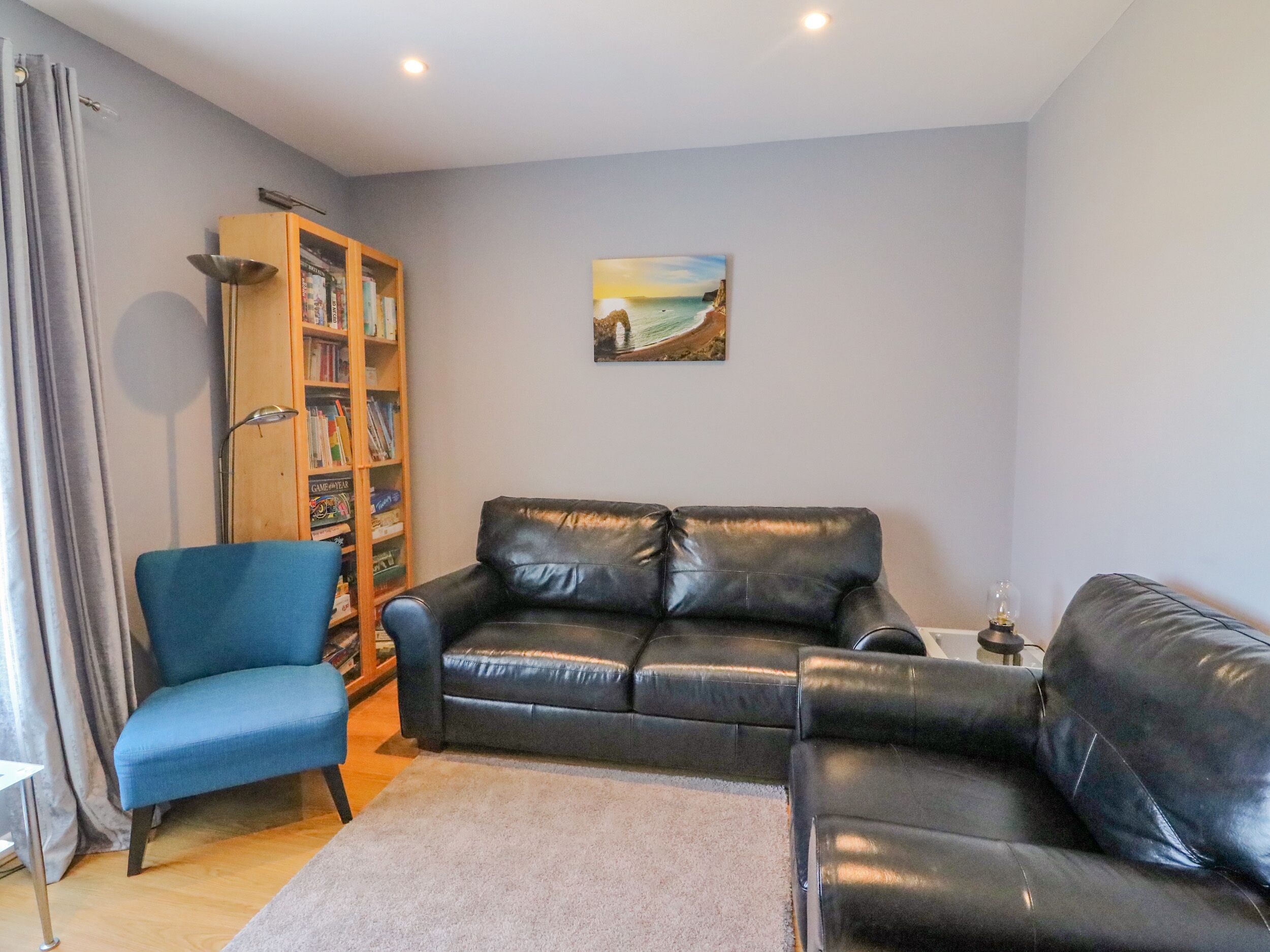 A living room with a blue armchair, a black leather sofa, a bookshelf, and a painting on the wall.
