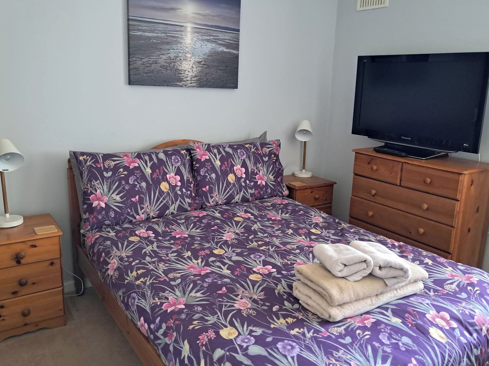 Un dormitorio con cama, televisor, mesita de noche, lámpara y cómoda.