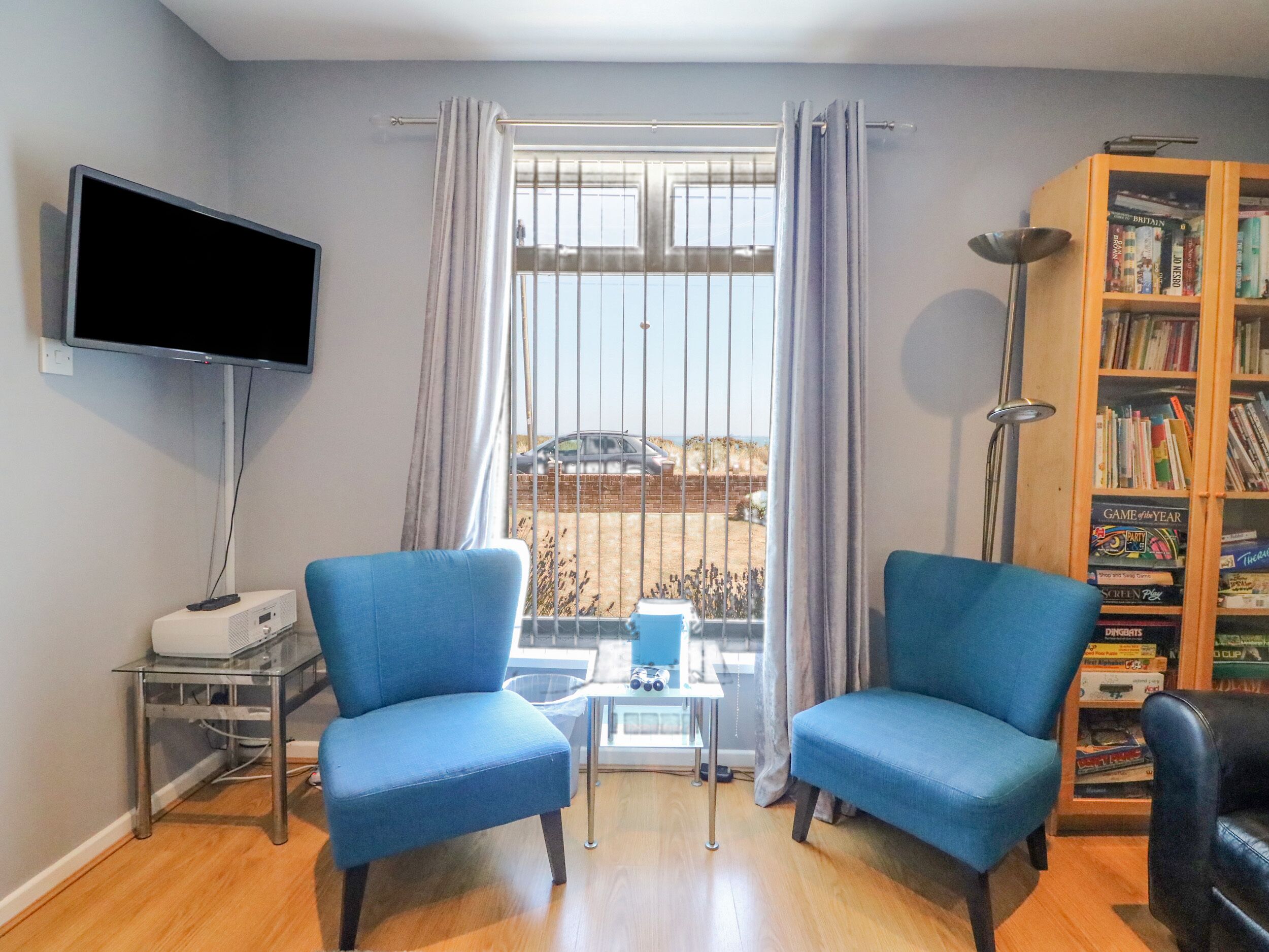 A living room with two blue armchairs, a flat-screen TV, a bookshelf, and a window with curtains.