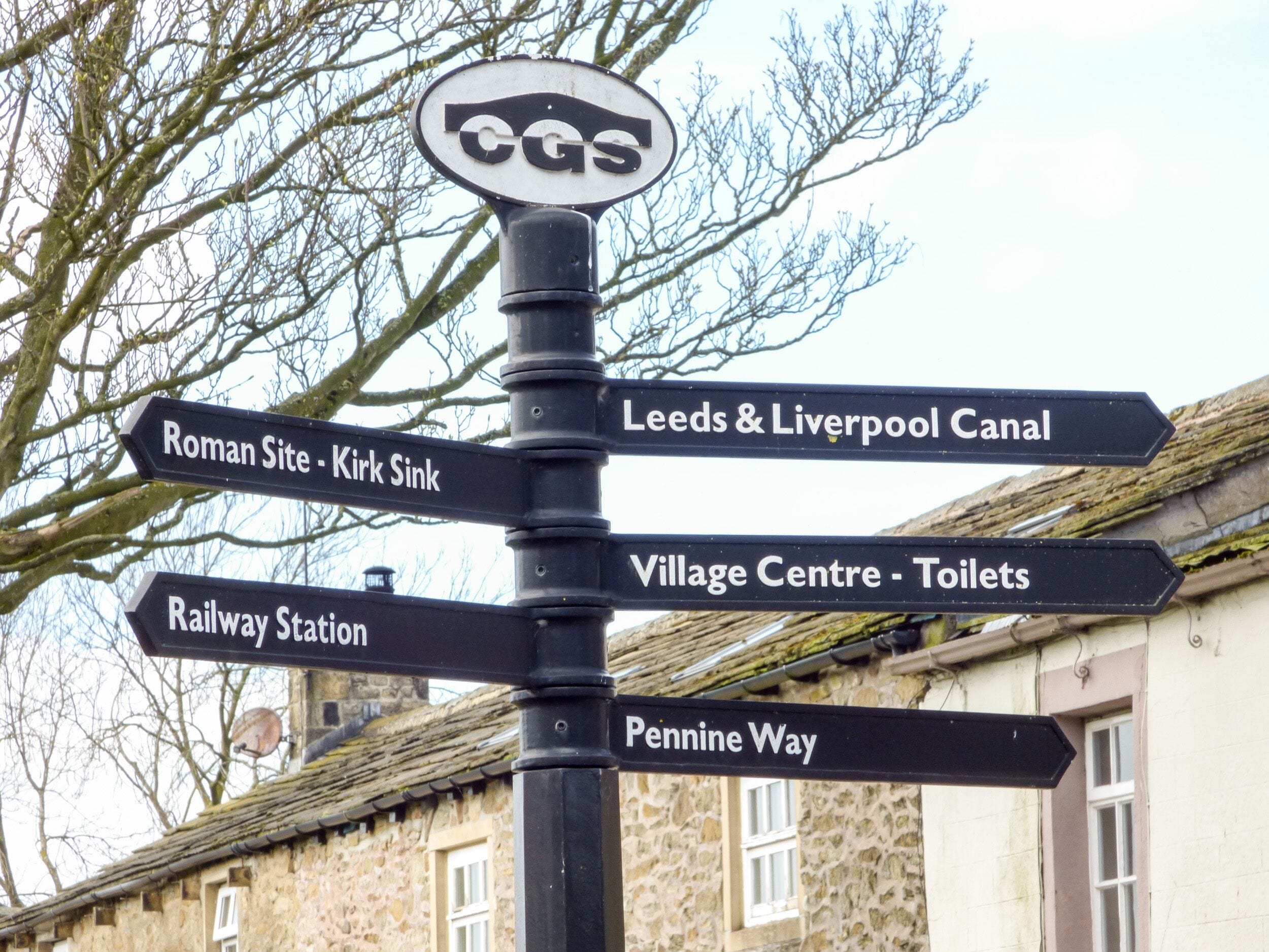 A directional signpost with multiple arrows pointing towards various destinations such as Roman Site, Kirk Sink, Leeds & Liverpool Canal, Village Centre, Railway Station, and Pennine Way.