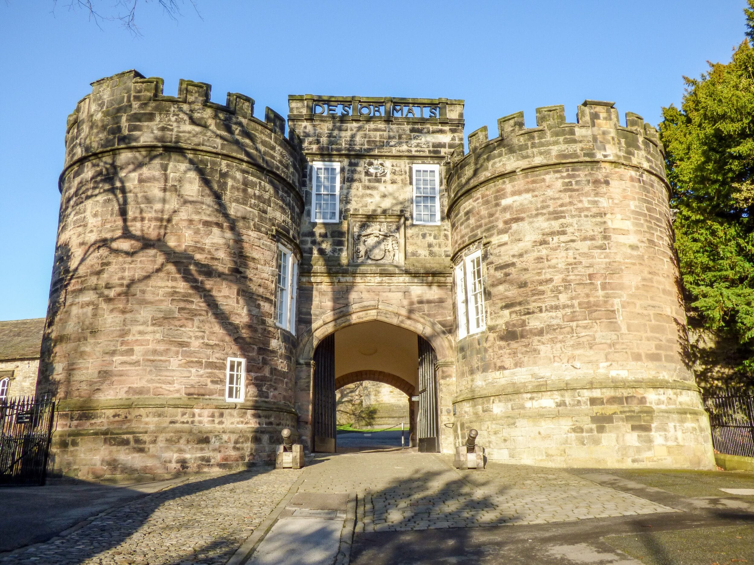 A historic stone castle entrance with a sign reading "DE'S OF MATS".