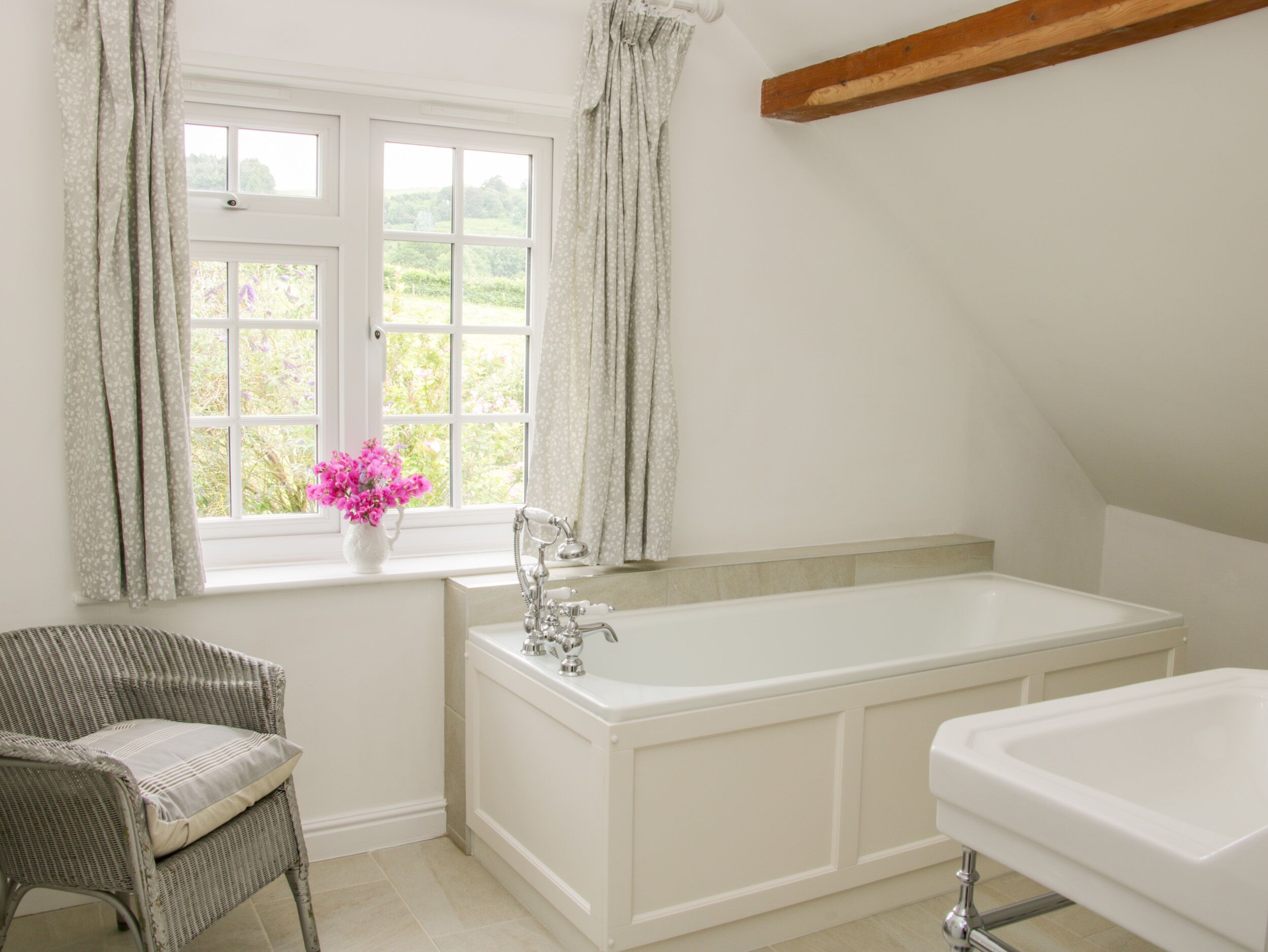 A bathroom with a bathtub, a window with curtains, and a wicker chair.