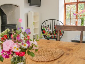 A cozy living room with a fireplace, a wooden table with a woven placemat, a black chair with colorful cushions, and a window with a view of greenery.