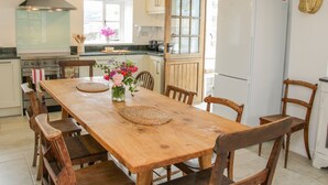 A wooden dining table with chairs, a kitchen with a stove, oven, and refrigerator, and a window with flowers.