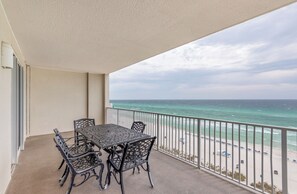 Un balcon avec une table et des chaises, donnant sur une plage, avec une rambarde et une vue sur l’océan.