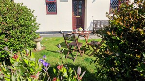 Uma casa de campo branca com porta vermelha, um pequeno jardim e um banco de madeira.