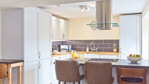 A modern kitchen with white cabinets, a glass backsplash, and a stainless steel range hood. There are two bar stools and a breakfast setup on the island. - Harley House (Broughton in Furness)