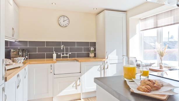 A modern kitchen with white cabinets, a black tiled backsplash, a clock, and a breakfast setup with croissants and orange juice. - Harley House (Broughton in Furness)