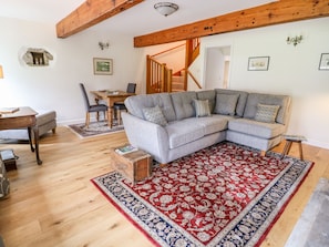 Ein gemütliches Wohnzimmer mit einem grauen Ecksofa, Holzboden, einem Teppich mit rotem und blauem Muster, einem Couchtisch aus Holz und einem Essbereich mit Holztisch und Stühlen.