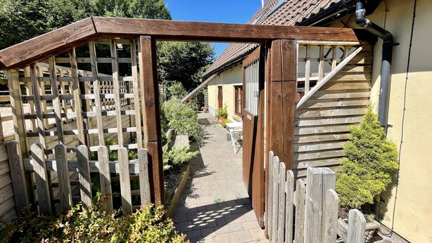 Ein schmaler Holzweg führt zu einem Haus mit Ziegeldach und einem Holzzaun an der Seite.