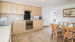 A modern kitchen with wooden cabinets, a built-in oven, a microwave, and a dining area with a table and chairs. - The Old Cook House (Penrith)