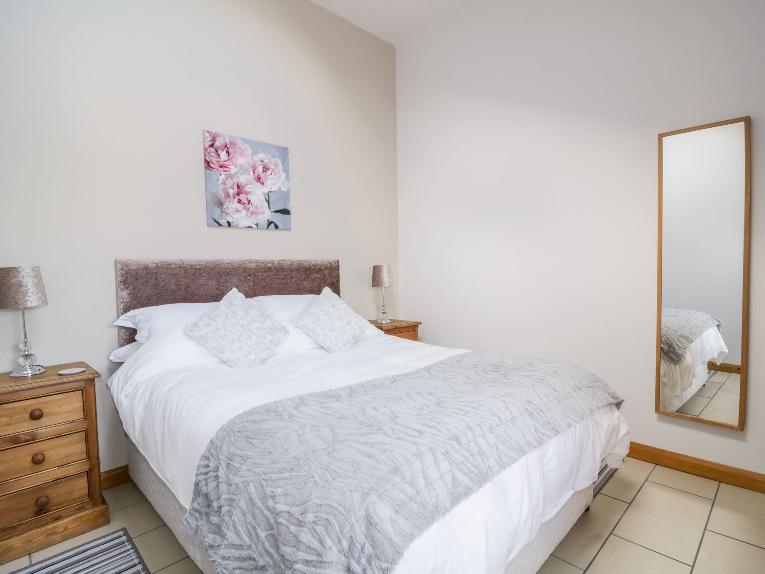 A neatly arranged bedroom with a bed, bedside tables, a lamp, a mirror, and a painting on the wall.