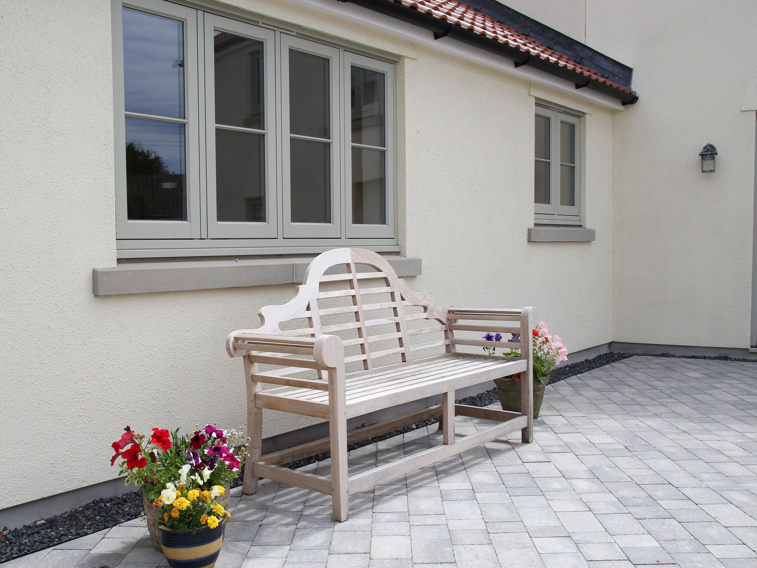 A wooden bench with a backrest and armrests, positioned on a paved patio area with a white wall and windows in the background.