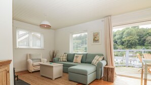 Un salon chaleureux avec un canapé vert, un fauteuil blanc et une table basse en bois. Dehors, il y a une terrasse en bois avec des chaises.