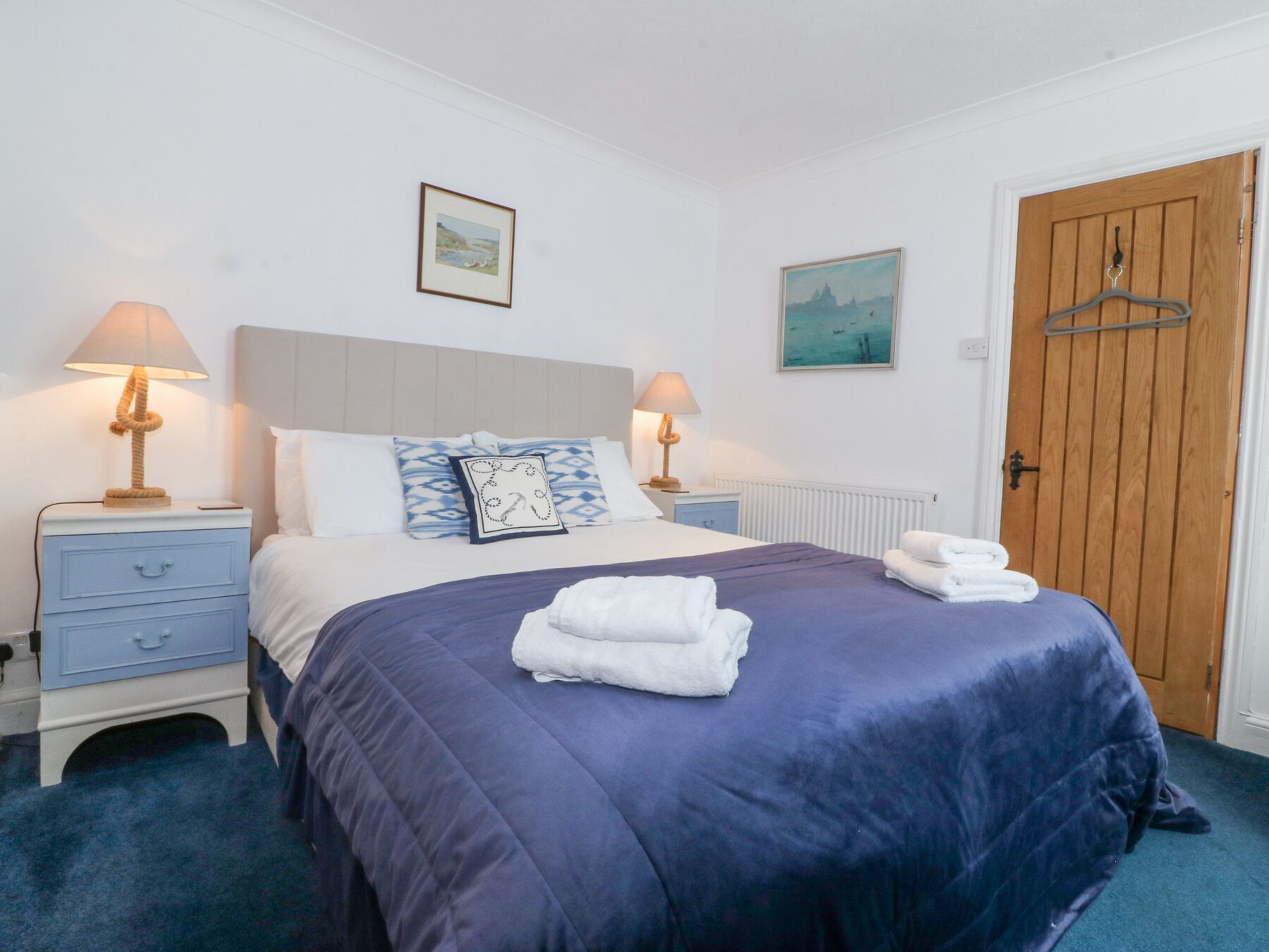 A neatly made bed with a blue bedspread and two pillows, flanked by bedside tables with lamps, a wooden door, and a wall-mounted painting.