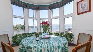 A corner dining area with a bay window overlooking a body of water, a table set with a bottle and glasses, and a framed picture on the wall. - Bay View Apartment (Porthmadog)