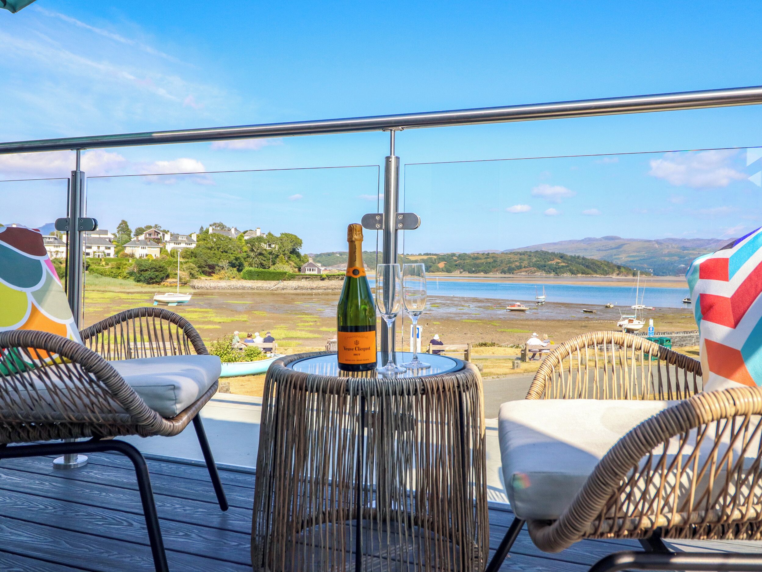 A balcony with wicker chairs, a bottle of champagne, and a view of a beach with boats.