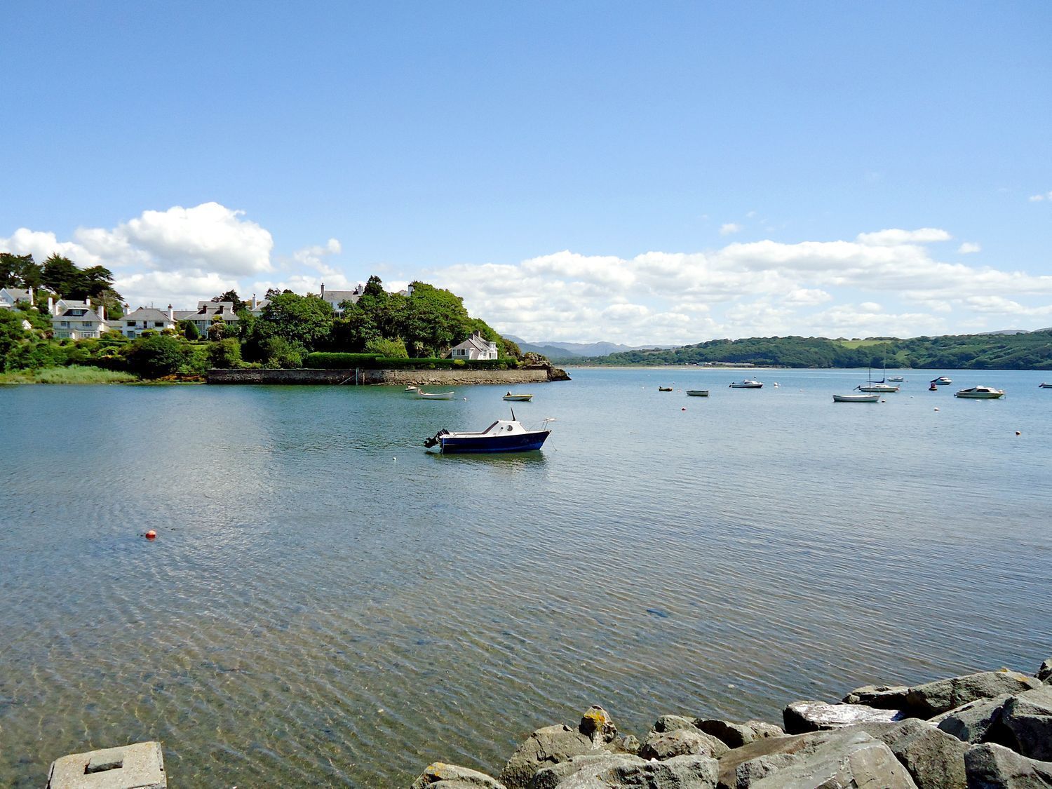 Um lago tranquilo com barcos, uma margem rochosa e casas ao fundo.