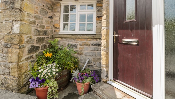 A stone building with a dark wooden door, a white window, and potted plants in front. - Coquet View Cottage (Morpeth)