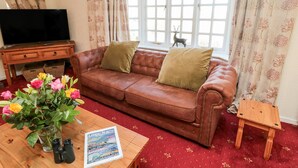 A living room with a brown leather sofa, a wooden coffee table with a magazine, a TV on a wooden stand, and a window with curtains. - Coquet View Cottage (Morpeth)