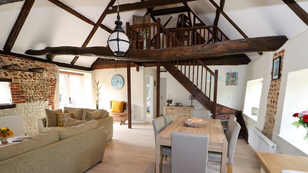 Ein geräumiges Loft mit freiliegenden Holzbalken, einem gemütlichen Wohnbereich mit Sofa und Esstisch sowie einer Treppe zu einer oberen Ebene.