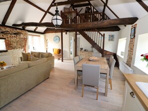 Een ruime loft met zichtbare houten balken, een gezellig woongedeelte met een bank en een eettafel, en een trap naar een hoger niveau.
