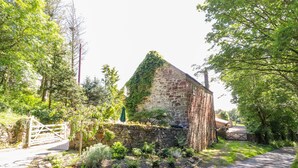 A rustic stone building with a steeply pitched roof, surrounded by lush greenery and a gravel path. - The Old Coach House (Mold)