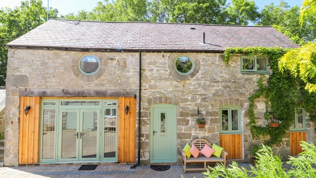 A stone building with two wooden doors and windows, a bench with cushions, and a potted plant. - The Old Coach House (Mold)