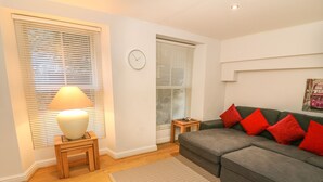 A modern living room with a grey sofa, red cushions, a wooden side table with a lamp, and a patterned rug. - Flat 2 Galloway House (Kendal)
