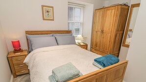 A bedroom with a wooden bed, a nightstand, a lamp, a wardrobe, and a window with blinds. - Flat 2 Galloway House (Kendal)