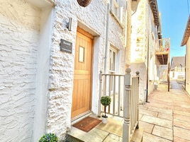 A narrow street with a white building featuring a wooden door and a small porch with a potted plant.