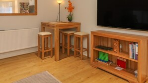 A cozy living room with a wooden table, two stools, a bookshelf, and a flat-screen TV. - Flat 2 Galloway House (Kendal)