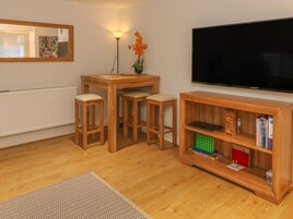 A cozy living room with a wooden table, two stools, a bookshelf, and a flat-screen TV.