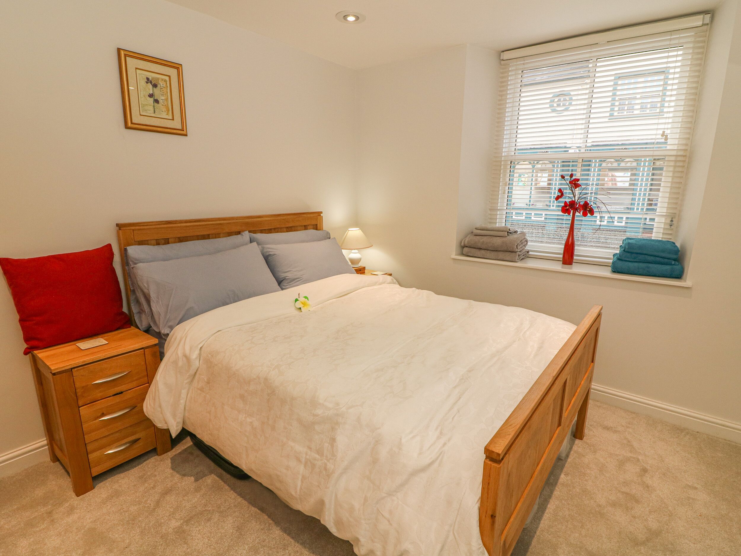 A neatly made bed with a wooden headboard, a nightstand with drawers, a bedside lamp, and a window with blinds and towels on the sill.