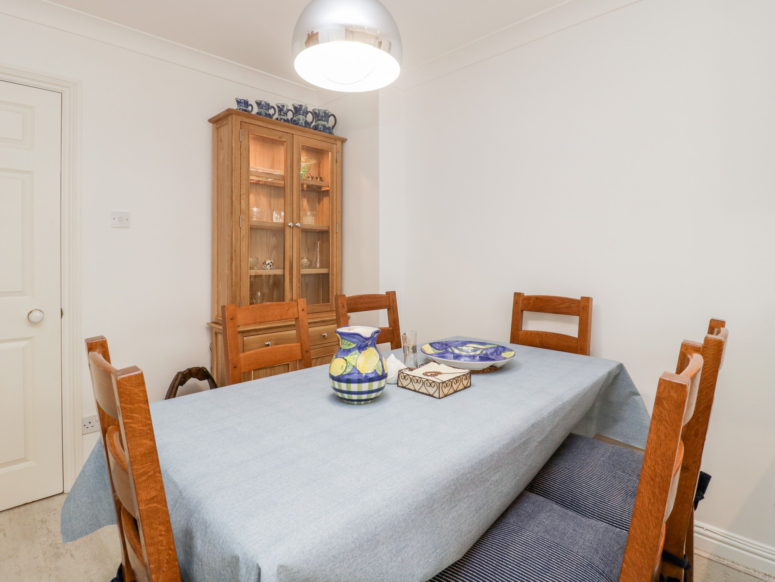 A dining area with a table set for four, wooden chairs, a cabinet with glass doors, and a ceiling light.