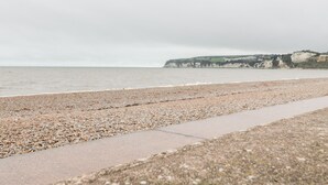 A pebbled beach with a concrete path leading to the water, and cliffs in the background. - 14 Lyme Mews (Seaton)
