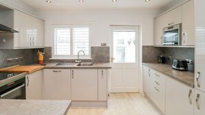 A modern kitchen with white cabinets, a built-in oven, a sink, and a window with blinds. - 14 Lyme Mews (Seaton)